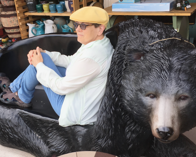 Short white woman with thick glasses, yellow Kongol flat cap, sitting back against the resin head of a black bear