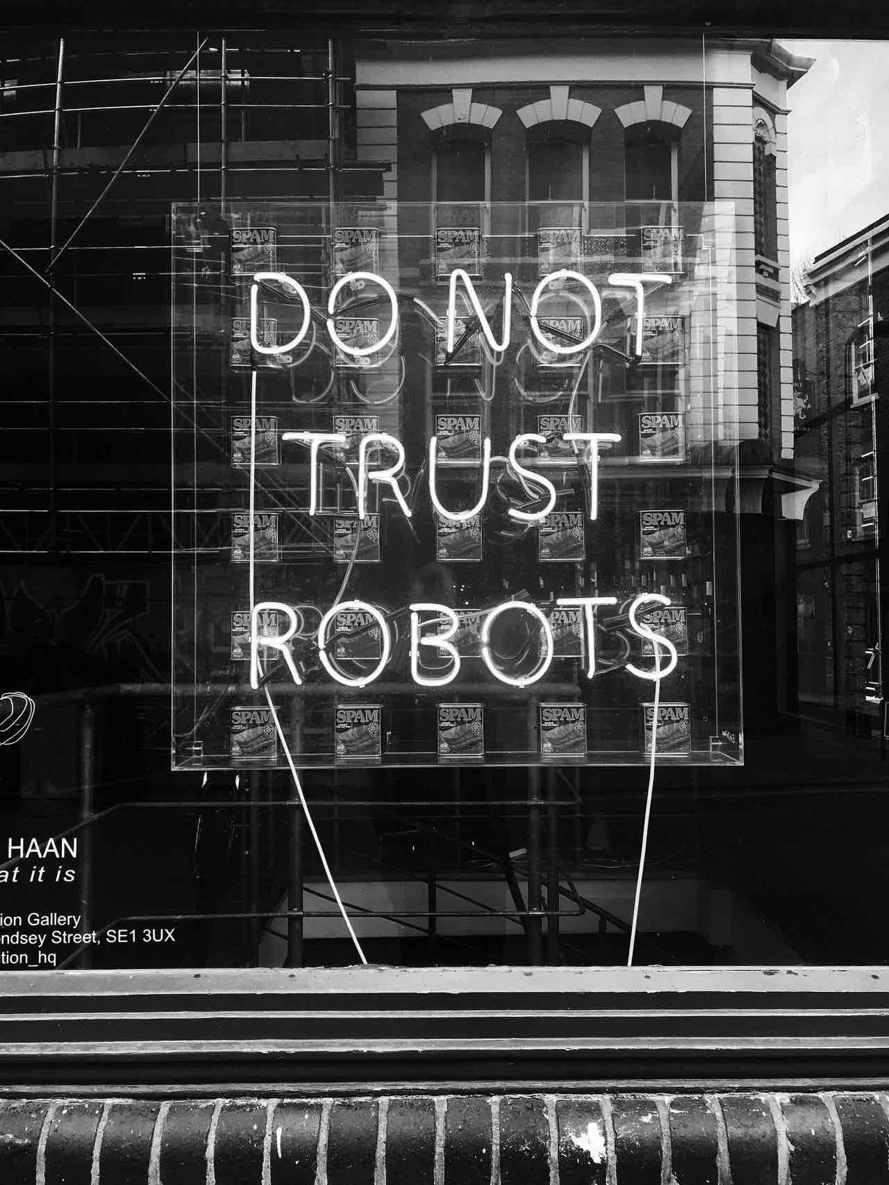 A black and white photo of a neon sign that says “Do Not Trust Robots” with cans of Spam behind it.