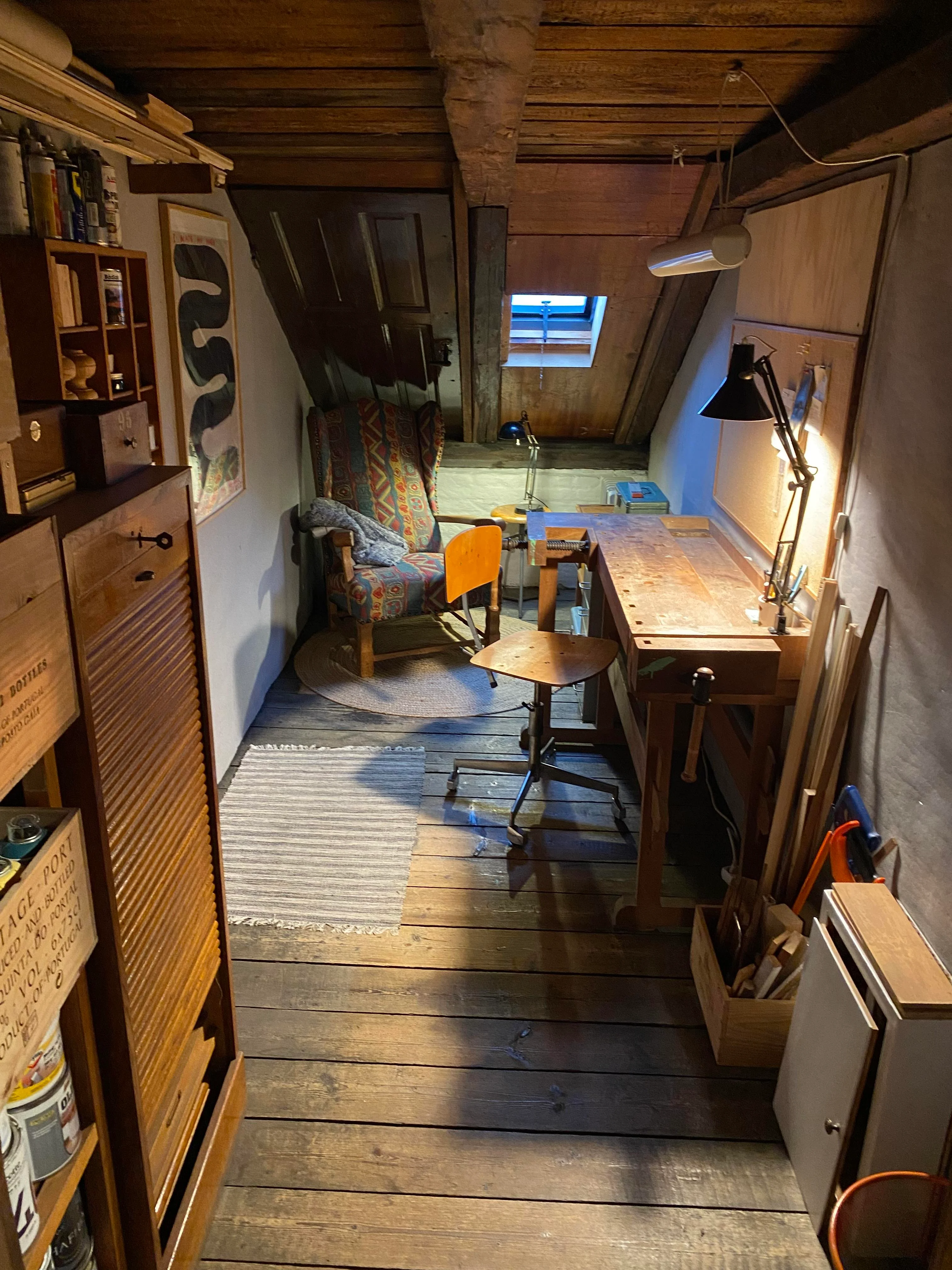 A narrow attic workspace with a workbench, comfy chair, shelves, a very small window, and lots of wood.