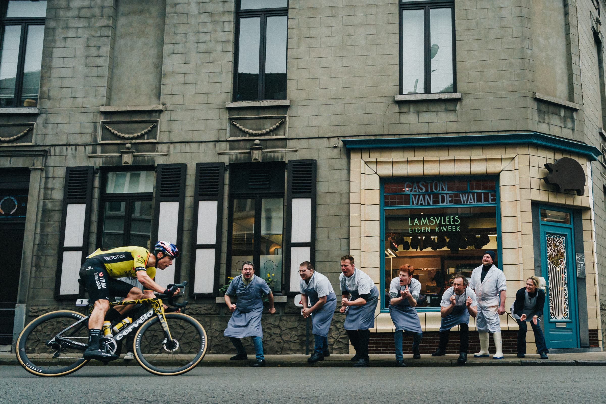 A bicyclist races through a city. A line of butchers cheer the cyclist on as they pass their shop.